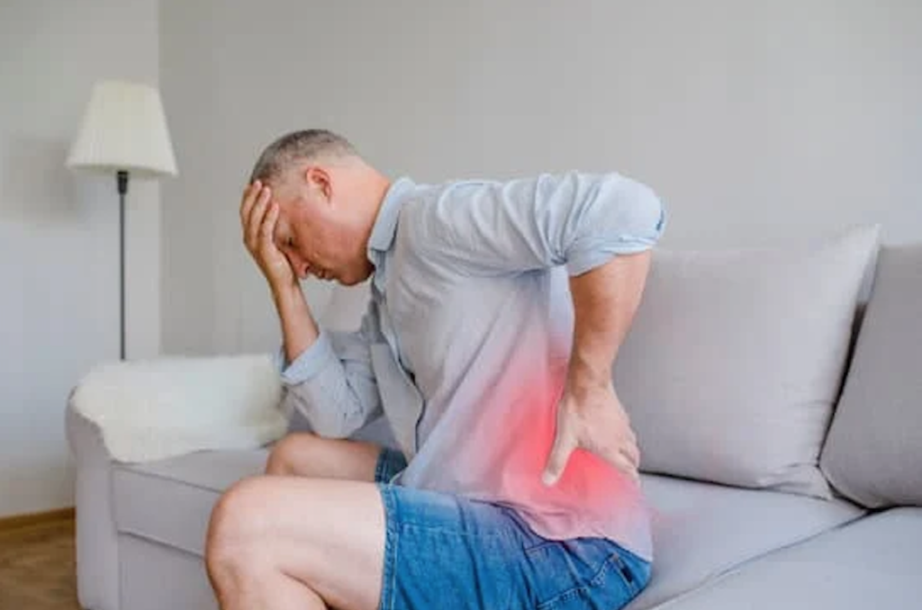 Man seated on a couch holding his lower back during a moment of discomfort at home